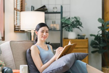 Güzel, genç, gülümseyen ve neşeli bir kadın oturma odasında bir kanepede otururken kitap okuyor.