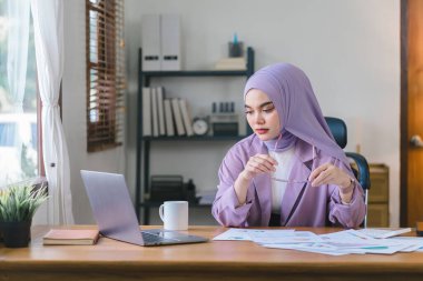 Memleketten çalışan, mor tesettürlü Müslüman bir kadın dizüstü bilgisayarını kullanırken endişeli, yorgun ve bunalmış görünüyor.