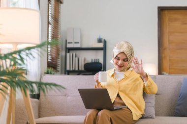 Çekici ve mutlu Müslüman milenyum iş kadını oturma odasındaki koltukta oturan tesettürlü bir iş kadını bilgisayarla görüntülü konuşma yapıyor ve el sallıyor.