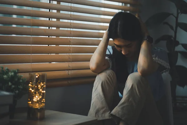 Sad and depressed young woman sitting nere window in the living room, looking outside with a sad expression, conveying feelings of exhaustion, loneliness, and unhappiness