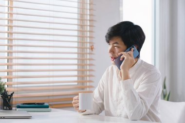 Milenyum Asyalı erkek serbest çalışan akıllı telefonuyla ev ofisinden bilgisayarıyla uzaktan çalışırken konuşuyor.