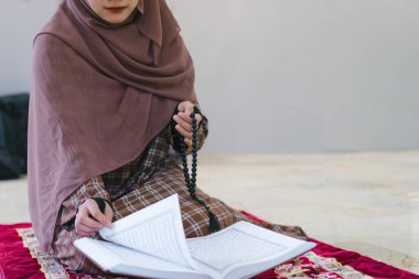 Kırpılmış bir görüntü, Müslüman bir kadının uzun bir İslami tesettür takıp kırmızı bir paspasın yanında duran Kur 'an okuduğunu gösteriyor. Seçici odak ve kopyalama alanı.