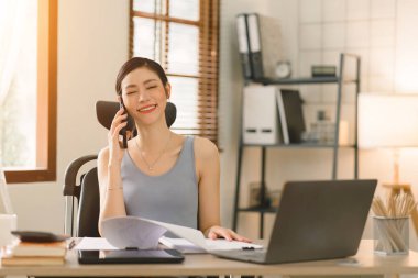 Yeni yetme bir iş kadını akıllı telefonuyla konuşuyor ve evinde otururken dizüstü bilgisayarı üzerinde çalışıyor.