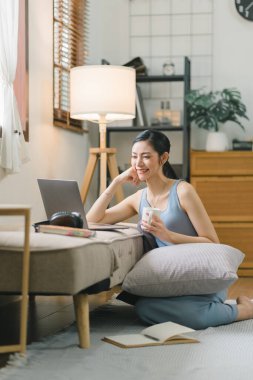 Millennial Asyalı kız oturma odasında kanepede oturup dizüstü bilgisayar başında ders çalışıyor. Yoğunlaştırılmış genç kadın bilgisayarda çalışıyor ve not defterine yazıyor, evde online kurs ya da eğitim görüyor.