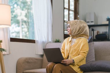 Ev ofisinden çalışırken oturma odasında bir kanepede oturan, sarı tesettürlü, çarpıcı bir Müslüman iş kadını. Pencerenin dışına bak.