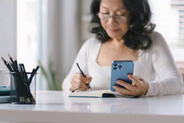 Uzaktan kumandayla akıllı telefondan ev telefonuyla çalışan çekici kıdemli iş kadını. Gülümseyen 60 'ların orta yaşlı iş kadını akıllı telefon kullanıyor. Webinar izliyor ve deftere yazı yazıyor.