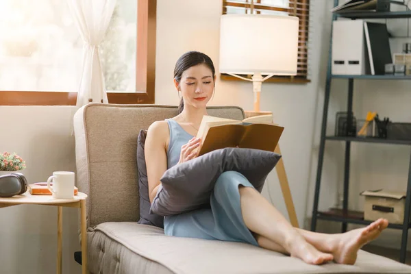 stock image A beautiful, young, smiling and cheerful woman is reading a book while sitting on a couch in the living room