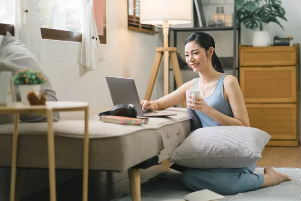 stock image Millennial Asian girl sits on sofa in living room, studying on laptop making notes.The concentrated young woman works on computer and writes in her notebook, taking online course or training at home