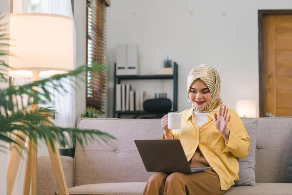 Müslüman bir kadın, iş arkadaşlarıyla telekonferansta bilgisayarında bir video görüşmesi yaparken görülüyor. Oturma odasında bir kanepede oturuyor ve kameraya doğru gülümseyip el kol hareketi yaparken görülebilir.
