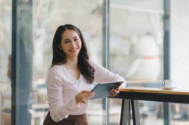 Gülümseyen, genç, güzel, profesyonel ve kendine güvenen Asyalı bir iş kadınının dijital tableti kullanarak ortak çalışma alanındaki satış verilerini analiz eden bir portresi.
