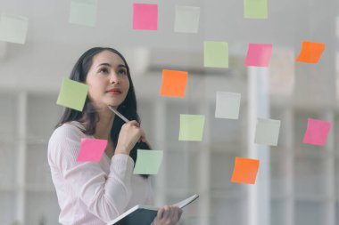Asyalı profesyonel bir iş kadını renkli yapışkan notlar yazıp yaratıcı ofisteki cam bir pencereye yapıştırıyor. İş planlaması ve beyin fırtınası stratejisi kavramı