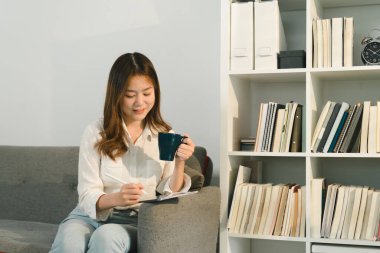 Büyüleyici Asyalı kadın evdeki kanepede dinlenirken dijital tabletini kullanıyor.