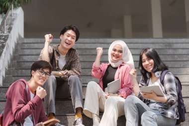 Bir grup çok ırklı ve mutlu genç, kutlama için ellerini kaldırıyor. Birlikte eğleniyorlar. Kampüs merdivenlerinde oturup, farklı arkadaşlar olarak birlik olduklarını gösteriyorlar.