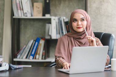 Güzel bir Müslüman milenyum Asyalı iş kadını ev ofisinden uzaktan çalışan kahverengi bir başörtüsü takıyor.
