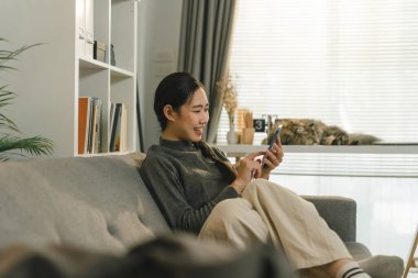 Çekici Asyalı kadın evdeki kanepede dinlenirken akıllı telefonunu kullanıyor.