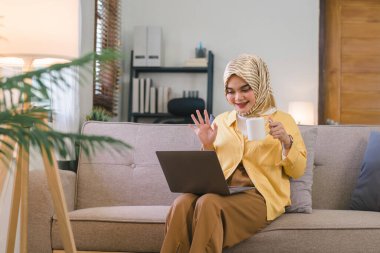Müslüman bir kadın, iş arkadaşlarıyla telekonferansta bilgisayarında bir video görüşmesi yaparken görülüyor. Oturma odasında bir kanepede oturuyor ve kameraya doğru gülümseyip el kol hareketi yaparken görülebilir.