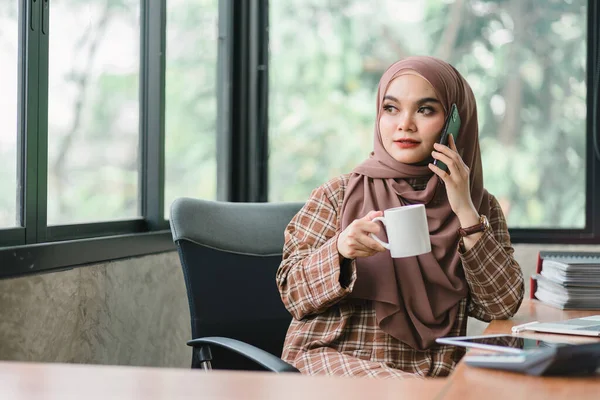 明るく楽しい100年に一度のアジア系ムスリム女性は オフィスでリラックスしながらコーヒーを飲みながらヘッドフォンで音楽を聴くのを楽しんでいます — ストック写真