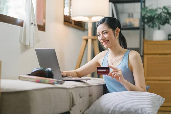 stock image A happy young woman is shown shopping online using her laptop and credit card at home