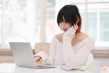 Genç bir Asyalı kadın rahat işyerinden internet üzerinden iletişim kurmak için bilgisayarını kullanıyor. Bu, uzaktan çalışma ve e-öğrenme kavramını betimler
