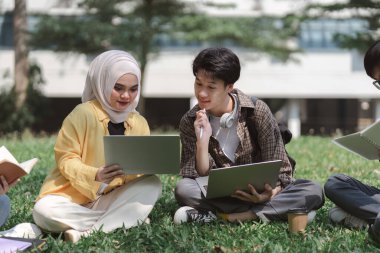 Asyalı üniversite öğrencilerinden oluşan çok ırklı bir grup kampüsün yakınındaki çimlerin üzerinde otururken birlikte ders çalışırken görülüyor.