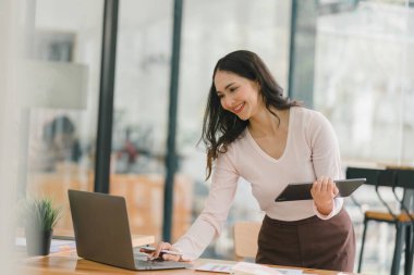 Gülümseyen, genç, güzel, profesyonel ve kendine güvenen Asyalı bir iş kadınının dijital tableti kullanarak ortak çalışma alanındaki satış verilerini analiz eden bir portresi.