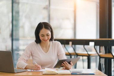 Gülümseyen, genç, güzel, profesyonel ve kendine güvenen Asyalı bir iş kadınının dijital tableti kullanarak ortak çalışma alanındaki satış verilerini analiz eden bir portresi.