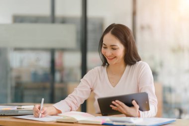 Gülümseyen, genç, güzel, profesyonel ve kendine güvenen Asyalı bir iş kadınının dijital tableti kullanarak ortak çalışma alanındaki satış verilerini analiz eden bir portresi.