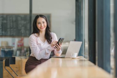Kafedeki güzel Asyalı iş kadını, akıllı telefon kullanıyor ve gülümsüyor.
