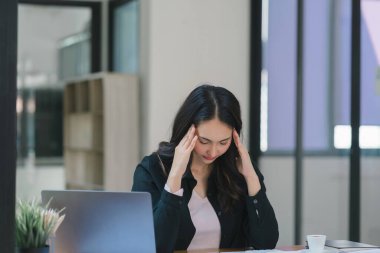 Stresli ve yorgun Asyalı iş kadını ofis masasında eli başının üstünde otururken görülüyor. Bu da yoğun bir iş günü geçirdiğini gösteriyor.