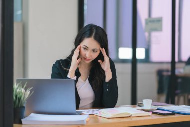 Stresli ve yorgun Asyalı iş kadını ofis masasında eli başının üstünde otururken görülüyor. Bu da yoğun bir iş günü geçirdiğini gösteriyor.