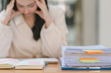 Stresli ve yorgun Asyalı iş kadını ofis masasında eli başının üstünde otururken görülüyor. Bu da yoğun bir iş günü geçirdiğini gösteriyor..