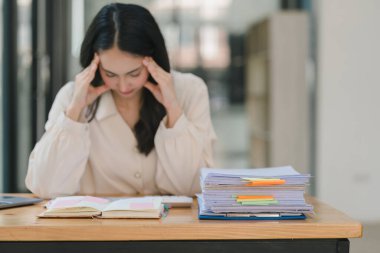 Stresli ve yorgun Asyalı iş kadını ofis masasında eli başının üstünde otururken görülüyor. Bu da yoğun bir iş günü geçirdiğini gösteriyor..