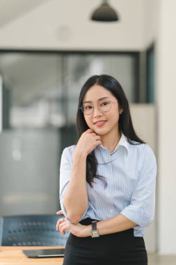 Modern bir ofiste duran gülümseyen, genç, güzel, profesyonel ve kendinden emin Asyalı bir iş kadınının portresi.