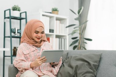 Evde tabletini kullanan, oturma odasındaki bir kanepede otururken sosyal ağlara mesaj atan ya da göz gezdiren Asyalı Müslüman bir kızın portresi.
