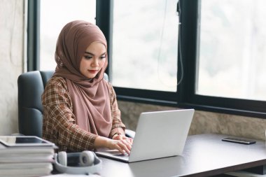 Kendine güvenen, güzel, profesyonel, bin yıllık Müslüman, kahverengi başörtüsü takan Asyalı bir iş kadını modern ofisten uzaktan çalışıyor.