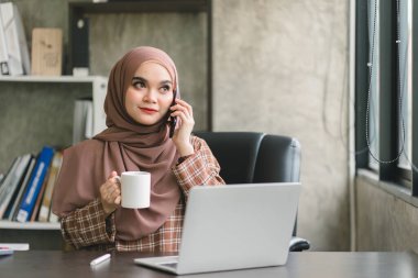 Güzel bir Müslüman milenyum Asyalı iş kadını ev ofisinden uzaktan çalışan kahverengi bir başörtüsü takıyor.