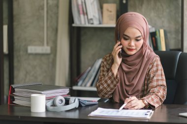 Kendine güvenen, güzel, profesyonel, bin yıllık Müslüman, kahverengi başörtüsü takan Asyalı bir iş kadını modern ofisten uzaktan çalışıyor.