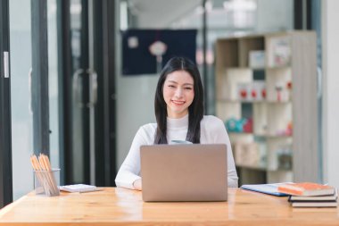 Başarılı ve güzel bir Asyalı iş kadını, sekreter, satış müdürü ya da iş danışmanı portresi, iş yerinde dizüstü bilgisayarla oturuyor, gülümsüyor ve günlük kıyafetlerini giyiyor.