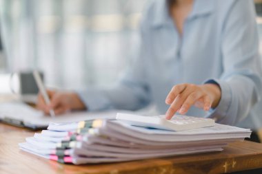 Sıradan giyinmiş profesyonel bir Asyalı iş kadını masasındaki rakamları hesaplamak için hesap makinesi kullanarak finansal verileri analiz ediyor.