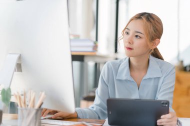Sıradan giysiler içinde odaklanmış ve profesyonel Asyalı bir iş kadını masaüstü bilgisayarı ve tabletindeki mali verileri analiz ediyor