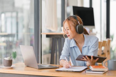 Kulaklık takan Asyalı iş kadını Webinar video kursunu izlerken not alıyor. İnternette okumak için e-öğrenim yoluyla ders dinleyen ciddi bir kız öğrenci.