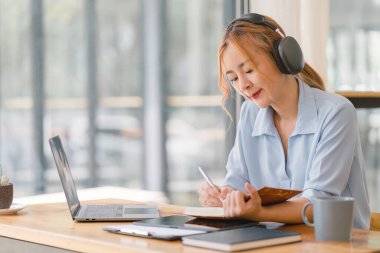 Kulaklık takan Asyalı iş kadını Webinar video kursunu izlerken not alıyor. İnternette okumak için e-öğrenim yoluyla ders dinleyen ciddi bir kız öğrenci.