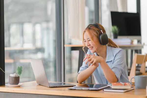 stock image Happy Asian businesswoman wearing headphones is taking notes in a notebook while watching a webinar video course. happy female student listening to the lecture to study online through e-learning