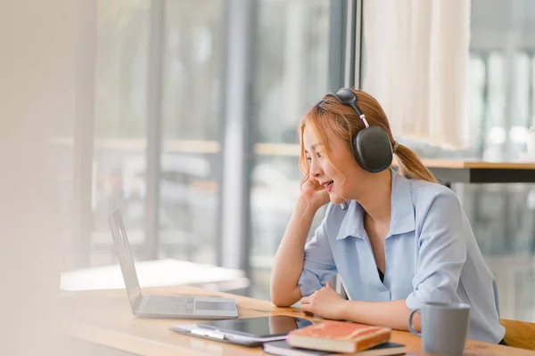 stock image Happy Asian businesswoman wearing headphones is taking notes in a notebook while watching a webinar video course. happy female student listening to the lecture to study online through e-learning