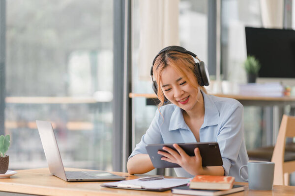 Focused Asian businesswoman wearing headphones is taking notes in a notebook while watching a webinar video course. Serious female student listening to the lecture to study online through e-learning