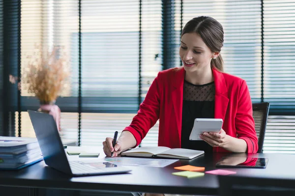 stock image Successful businesswoman in a stylish dress is sitting at a desk in a modern office, using a laptop computer. She appears to be a successful finance manager who is planning work projects