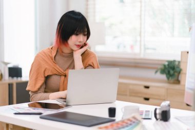 Gülümseyen, genç, güzel, profesyonel ve kendine güvenen, yaratıcı ve stil sahibi bir kadın tasarımcının portresi, ofisinde otururken bir tablet ve stilus kalem kullanıyor.