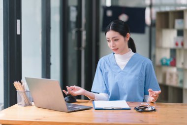 Kendine güvenen, güzel, profesyonel, bin yıllık bir kadın doktor ya da hemşire bir hastane danışma odasında bir masada otururken bilgisayarla çalışıyor.