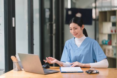 Kendine güvenen, güzel, profesyonel, bin yıllık bir kadın doktor ya da hemşire bir hastane danışma odasında bir masada otururken bilgisayarla çalışıyor.