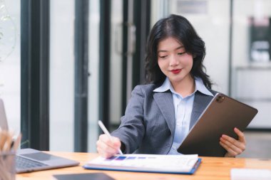 Smiling young Asian businesswoman leader holding a digital tablet, working in an office. Professional executive manager or saleswoman using corporate technology, confidently
